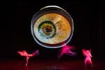 Large projected eyeball hovers above three dancers illuminated by red light onstage. Wayne McGregor's UniVerse: A Dark Crystal Odyssey, Company Wayne McGregor, Royal Opera House, London (2023), Photo credit/ Andrej Uspenski