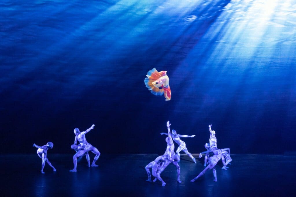 Company Wayne McGregor dancers in lycra catsuits dance beneath a projection of a solitary goldfish swimming in the sea. Wayne McGregor UniVerse: A Dark Crystal Odyssey, ensemble 2, 2023, credit Andrej Uspenski