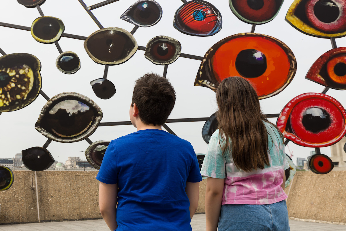 Two children looking at an artwork made up of cutouts of birds eyes. Installation view of Jenny Kendler, Dear Earth: Art and Hope in a Time of Crisis. Photo: Mark Blower. Courtesy the Hayward Gallery.