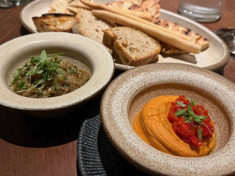 Two types of Greek dip with various breads; smoked aubergine dip and roast red pepper and feta dip topped with chopped sweet peppers.