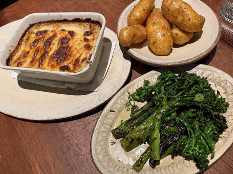 Three dishes on cream plates; an oven dish of moussaka with cheesy grilled topping, roast new potatoes, tenderstem broccoli