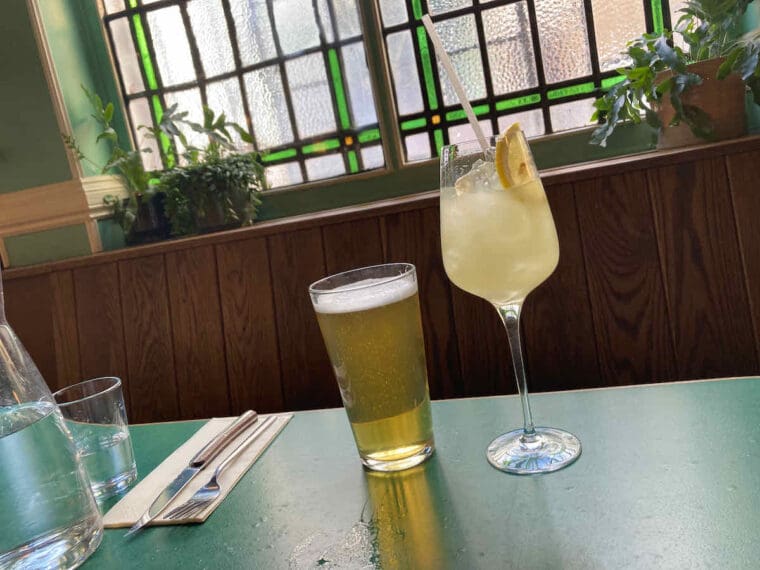 Two drinks on a green pub table with green stained glass window backdrop; a pint of lager alongside pale yellow Yuzu spritz cocktail in tall stem glass.