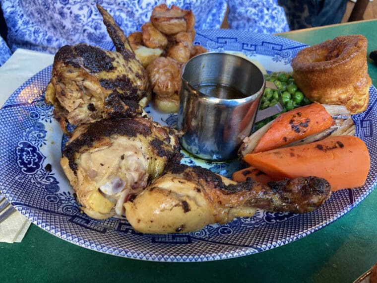 Sunday roast dinner on a blue and white willow pattern plate comprising soy roast chicken, carrots, Yorkshire pudding, peas and baby roast potatoes with a metal jug of self serve gravy in the centre.