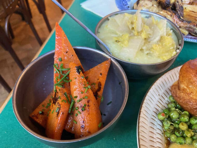 Two small metal bowls of vegetable side dishes; spears of chargrilled carrot with chive garnish and mashed potato with kimchi.
