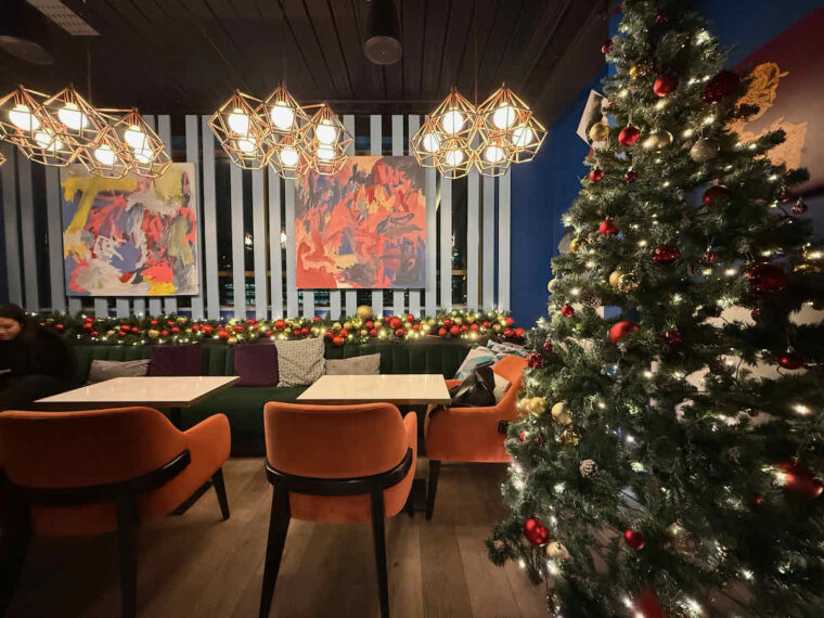 Interior shot of PocketSquare rooftop bar, Aldgate, East London; showing a large Christmas tree, orange velvet chairs, green banquet seating with various cushions, two square tables and three gold geometric metal light fittings, with six bare bulbs each, overhead. The back wall has two square abstract paintings.