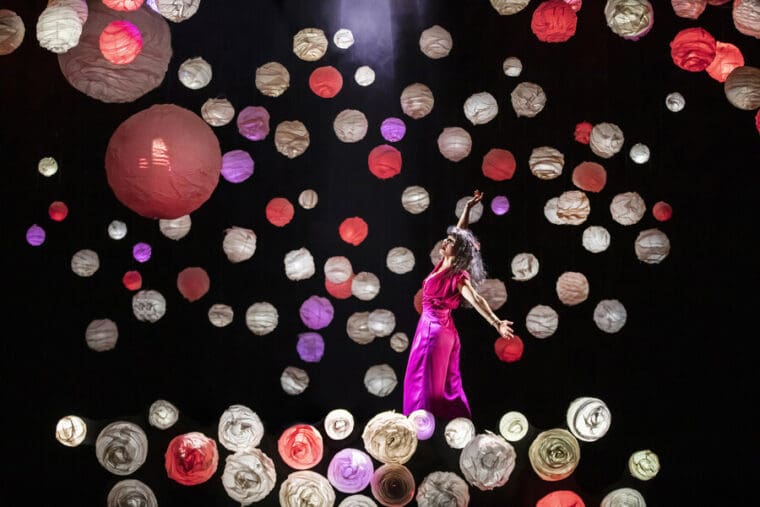 RSC A Midsummer Night's Dream Barbican pic credit Armani Ur-Rub; a dark background with Titania played by Sirine Saba standing with arms stretched up and behind her. She is dressed in a bright pink satin flowing jumpsuit and stood amidst many paper globes lit in shades of white, cream and pink giving a magical effect.