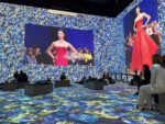 Vogue: Inventing the Runway, digital projection of blue flowers fill the walls and floor, two pictures fill the middle of each wall with a photo of a model in a red strapless dress. An audience are seated in the foreground.