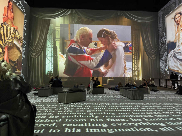 Vogue: Inventing the Runway at Lightroom, John Galliano dressed in union flag jacket holds a model by the shoulders, the floor of the room has printed text projected on it. 