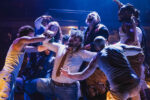 Animals attack Farmer Jones in Animal Farm at Stratford East; in dark dramatic light, a group of actors aggressively surround a man in casual check shirt and jeans. Photo credit Kirsten McTernan