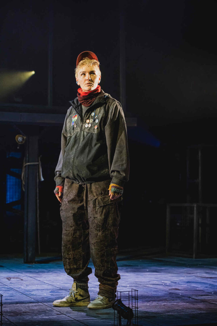 Em Prendergast as Milo in Animal Farm at Stratford East. They wear loose black clothes and a red baseball cap suggesting a bird's beak. Photo credit Kirsten McTernan