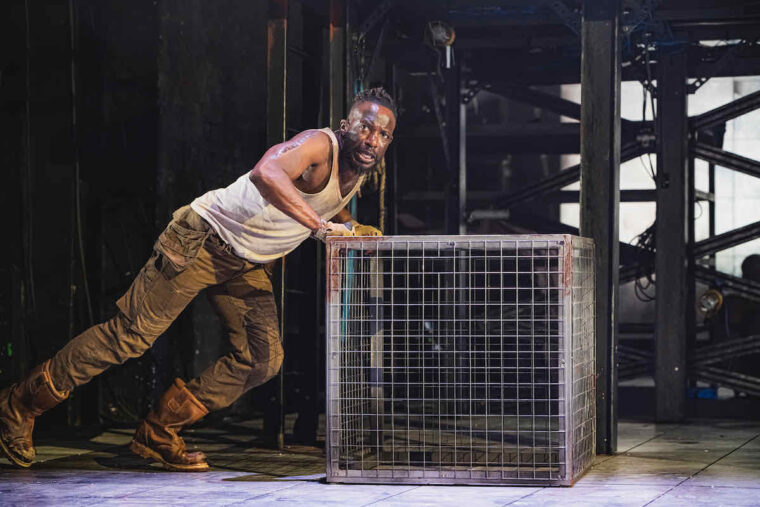 Gabriel Paul as Boxer in in Animal Farm at Stratford East; a muscular black man in vest and work trousers pushes a giant metal cage. Photo credit Kirsten McTernan
