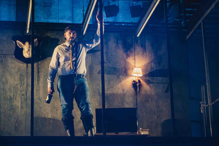 Kaya Ulasli as Farmer Jones in Animal Farm at Stratford East. A man drunkenly holding a beer bottle has his left arm raised; a pig's head is mounted on the industrial looking living room behind him. Photo credit Kirsten McTernan