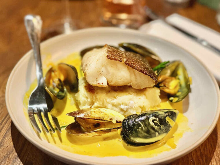 Wide bowl of roasted cod fillet on bed of mashed potato with six mussels surrounded by bright yellow mouclade sauce. As served at The Royal Forest, Epping Forest.