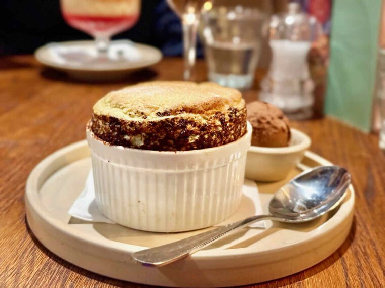 Pistachio soufflé dessert served at The Royal Forest, Epping Forest, Heartwood Inns. Golden brown pudding in a white ribbed ramekin, a spoon to the side, some chocolate ice cream behind. 