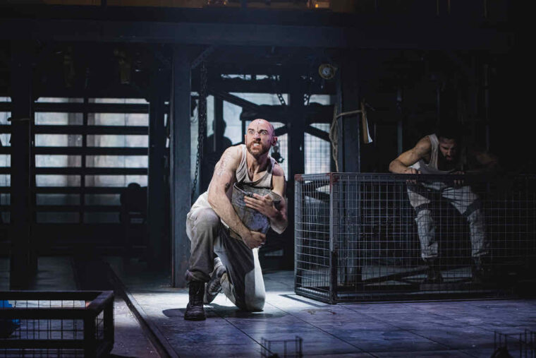 Tom Simper as Squealer the pig in Animal Farm at Stratford East; a white man with shaved head, white vest and jersey trousers, is poised on his knees clutching a metal bucket. The backdrop is dark and industrial looking. Photo credit Kirsten McTernan