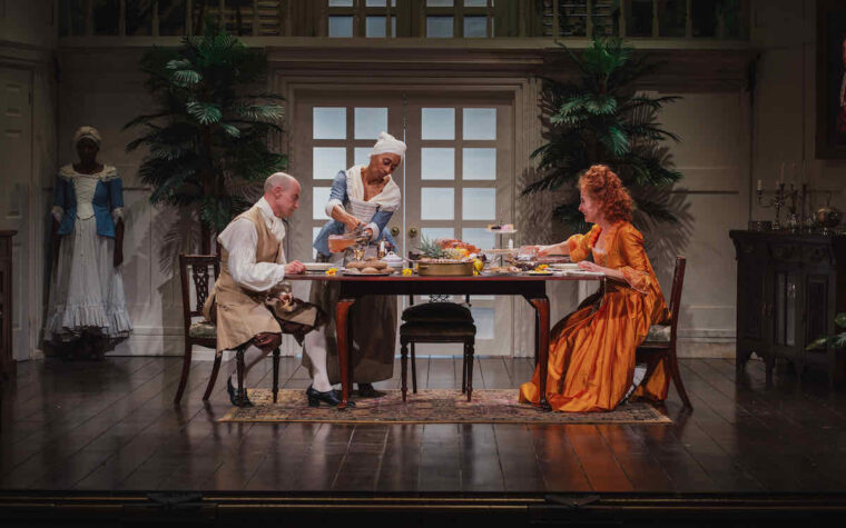 The cast of The Women of Llanrumney at Stratford East - white man and woman at dinner table being served by two black female slaves (c) Chuko Cribb