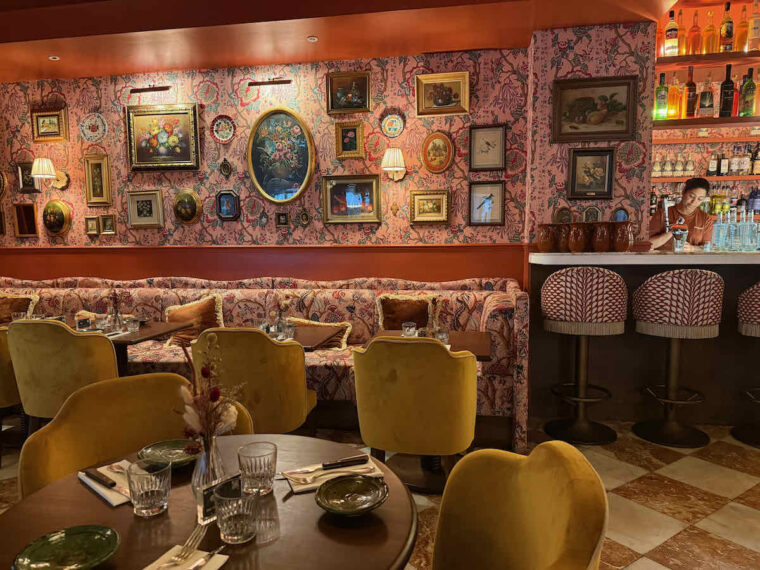 Tigermilk interior, Charing Cross Road, London. A gallery wall of framed pictures upon floral wallpaper with matching banquette seating and gold velvet chairs.