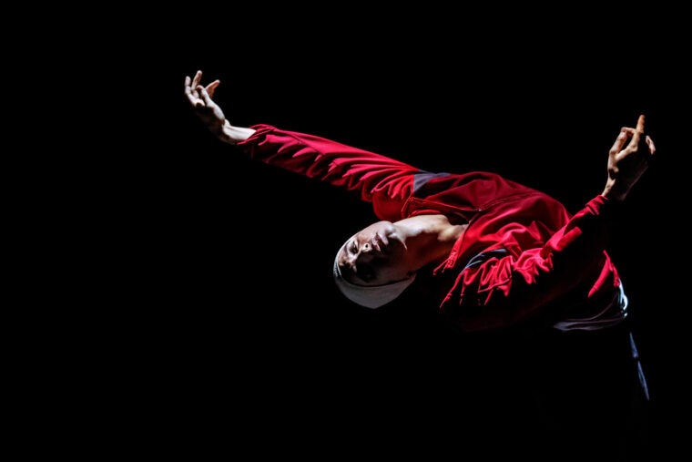 Dancer Daniel Proietto in red blouson and white woollen cap bends in Russell Maliphant's Afterlight