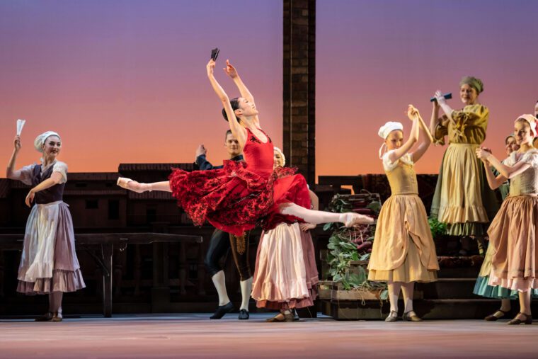 Kitri in a flouncy red dress and holding a fan, performs a split jete