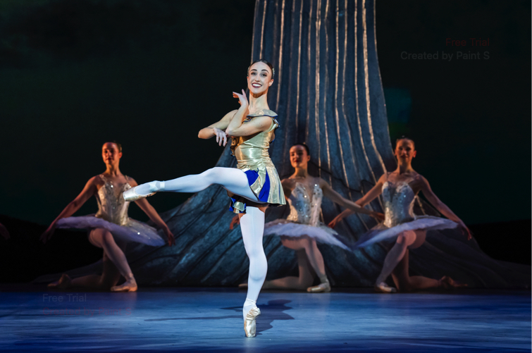 A woman in a gold and blue short costume stands on pointe with one leg in attitude devant in the Amor solo.