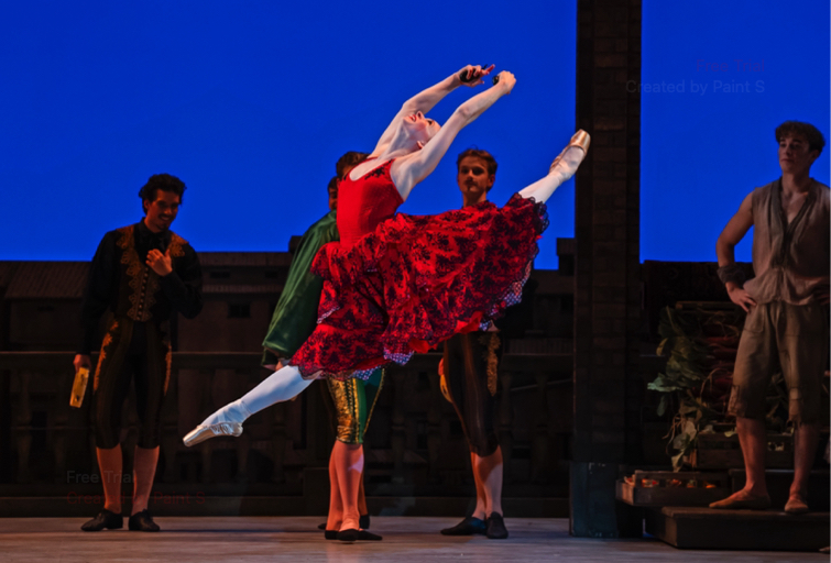 A dancer in a red dress with flounces, holding castanets performs a split jeté watched by teh villagers
