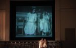 Two women sit on chairs synchronising their movement with that of a black and white image of themselves projected.on a high screen behind them
