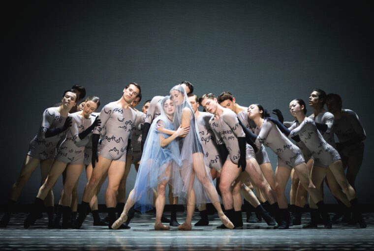 Two veiled women hold each other surrounded by an ensemble in grey leotards printed with Anne Lister's cypher