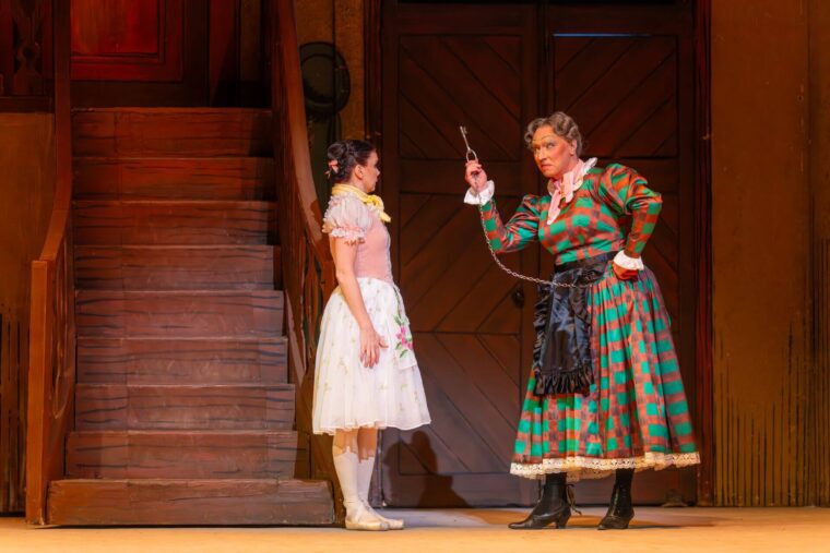 A woman in pink dress looks on as a stern looking older woman= in a checked green and brown long dress brandishes a door key.