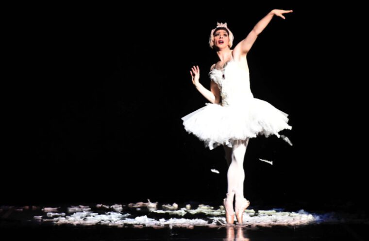 A ballerina on pointe in a white tutu moulting profusely all over the stage. Her right arm is stretched up, her face shows exaggerated suffering.