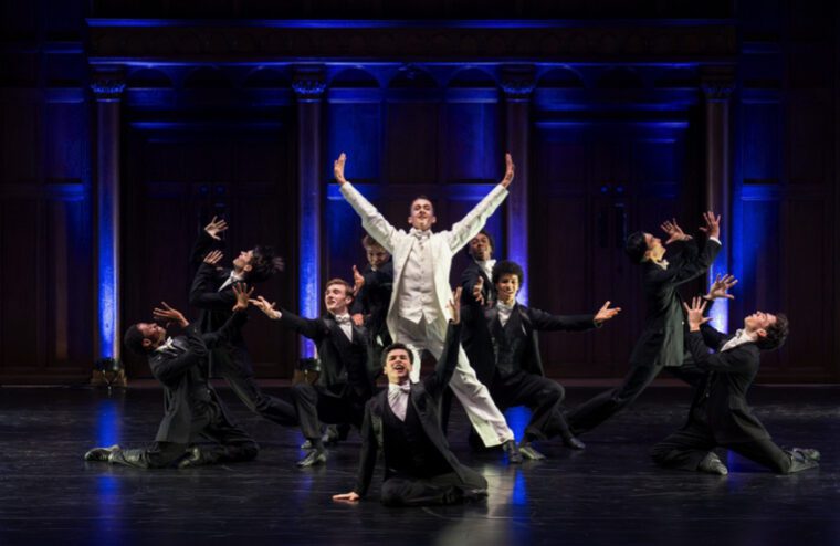 A group of men in bleck tuxedos posed around a man in white. Some are on their knees, all have arms raised