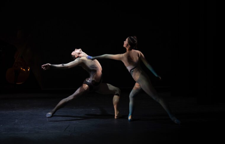 Next Generation Festival: two dancers in motley badysuits, the woman leans back with one leg stretched behind her, the man reaches out touching her shoulder.