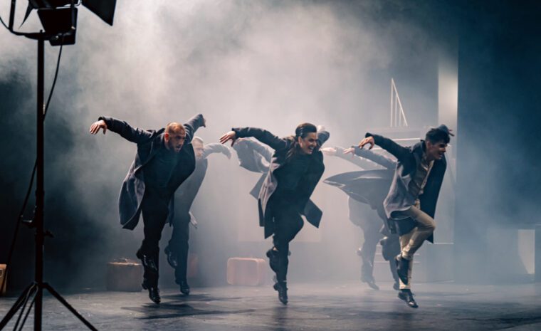 A line of three men in outdoor coats, do a tap dance in a smoky, moody atmosphere