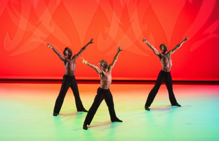 Against an orange background, three male dancers, their torsos bare. stand with legs apart and arms raised to heaven