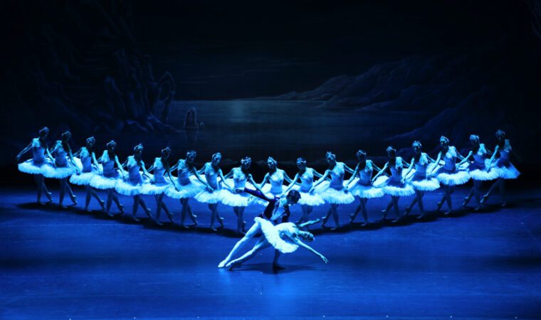 a scene from Swan Lake: Prince Siegfried holds Odette, who lies across his thigh. Behind them the corps of swans are perfectly aligned in an open V shape.
