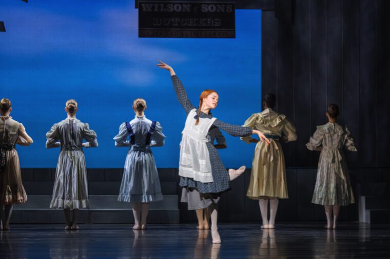A girl with red hair in plaits, wearing a grey dress and white smock stands on one leg with the other folded behind, right arm raised, left open to the side. Behind her the townswomen have her backs turned.