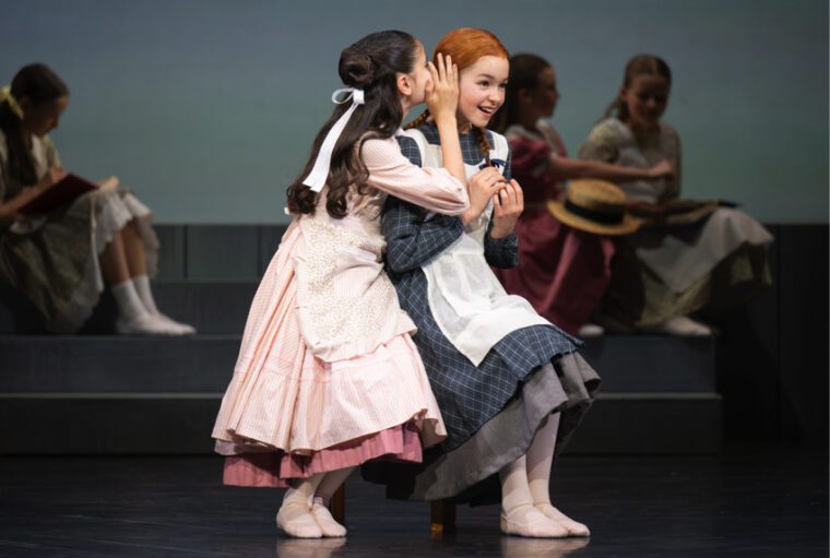 Anne of Green Gables: Anne's friend Diana, wearing a pink dress and white smock, whispers a secret in Anne's ear. Anne, in her grey dress and white smock, laughs.