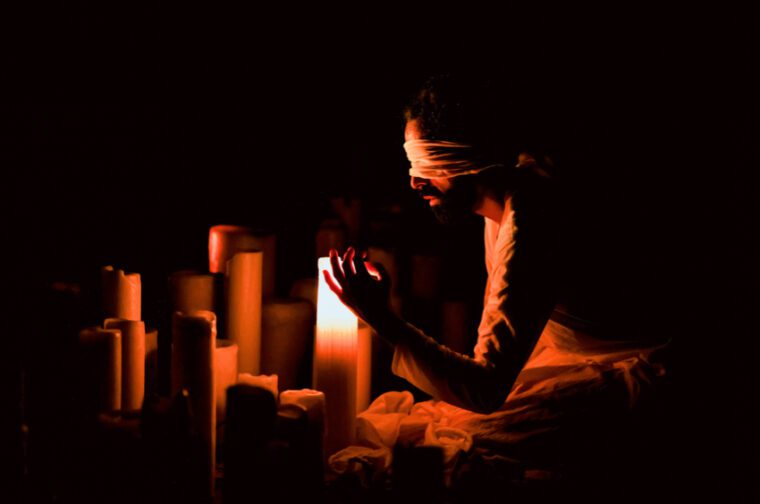 Songs of the Bulbul: the blindfold dancer sits before a line of short, thick candles. One candle is lit and illuminates his face