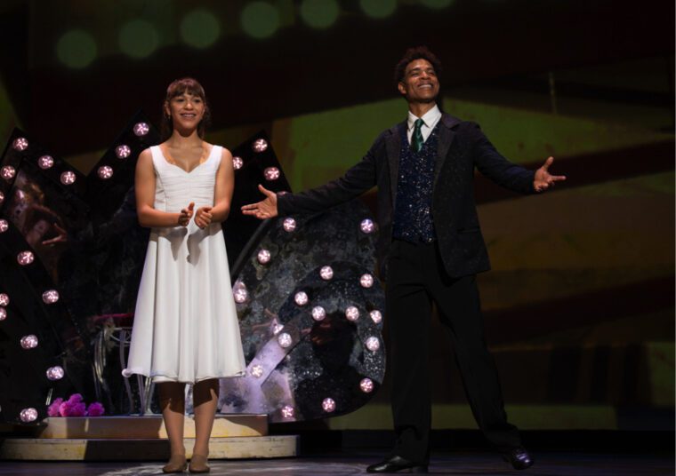 Nutcracker in Havana Suite: young Clara in a sleeveless white dress claps while Carlos Acosta in a three piece suite, stands to her left smiling with arms open
