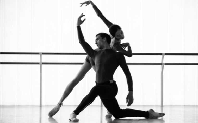 A black & white image of a studio rehearsal. A barre is visible at the back. The man kneels on his right knee, with the left leg bent in front. His right arm is raised, his head is titled over the right shoulder. The woman stands behind leaning on him, with the left leg stretched behind, left arm parallel to his right.