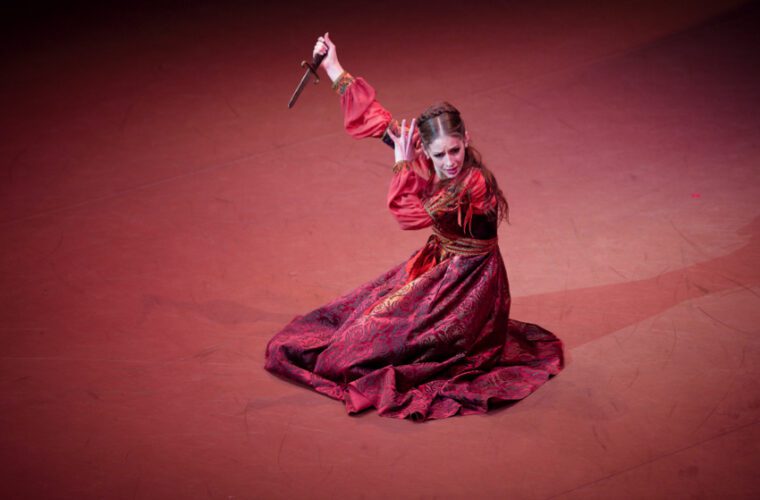 A dancer clad in a sumptuous red and gold brocade Renaissance gown kneels on the ground. Her right hand holds a dagger aloft, the left frames her grieving face.