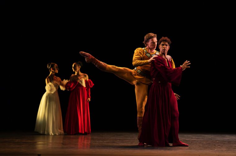 The Moor's Pavane: Max Maslen as Iago, dressed in a gold Renaissance inspired costume stands behind Carlos Acosta's Othello, dressed in dark red velved, whispering in his ear, Upstage Desdemona in white and Emilia in red talk together