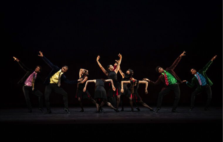 An ensemble scene from Rooster: The men in colourful shirts and ties have their arms raised. A central group of women surrounds on woman with both arms raised