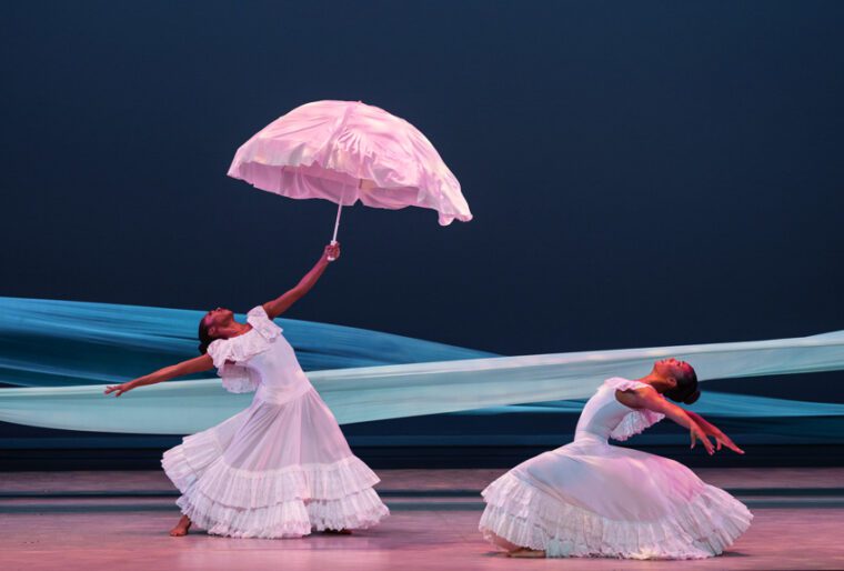 Alvin Ailey American Dance Theatre, Revelations. Against a dark blue background, two blue strips of cloth simulate a flowing river. Two women in ample, flouncy white dresses stand, one holding a white parasol, both in deep backbends.