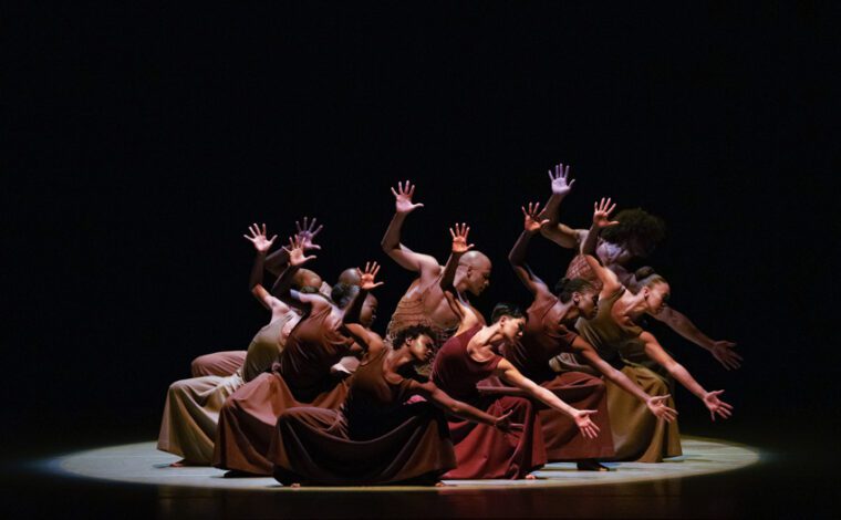 Alvin Ailey American Dance Theatre, Revelations. A cluster of dancers clad in deep earth tones, stand in deep knee bend leaning to our right, with the left arm stretch sideways, the right raise up, fingers splayed