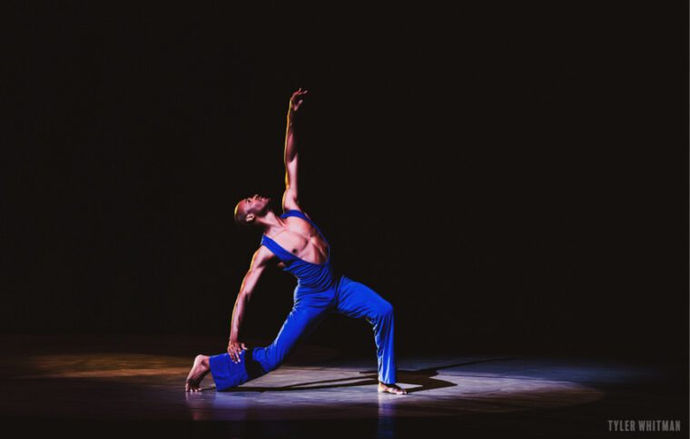 Alvin Ailey American Dance Theater, A Song For You. A man in a deep blue body suit with a plunging neck lline stands on a bent knee, his right leg behind, left arm and head lifted high.