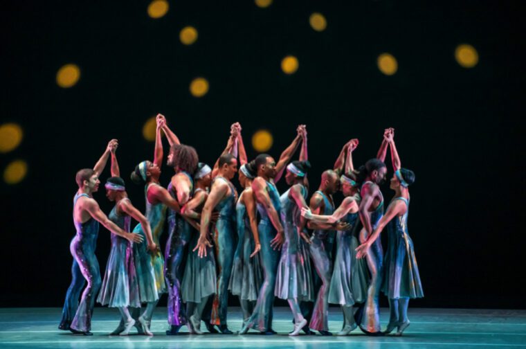Alvin Ailey American Dance Theater, Night Creature. Pairs of dancers in jazzy shimmering blue costumes stand in line, hands held above their heads