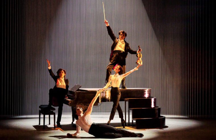 The McRae/Vassilev Project: the four performers, identically dressed in black, are grouped around a piano: the pianist sits at tke keyboard, the violist stands on the piano, McRae lies in the foreground holding the hand of Lamb who stands on pointe.