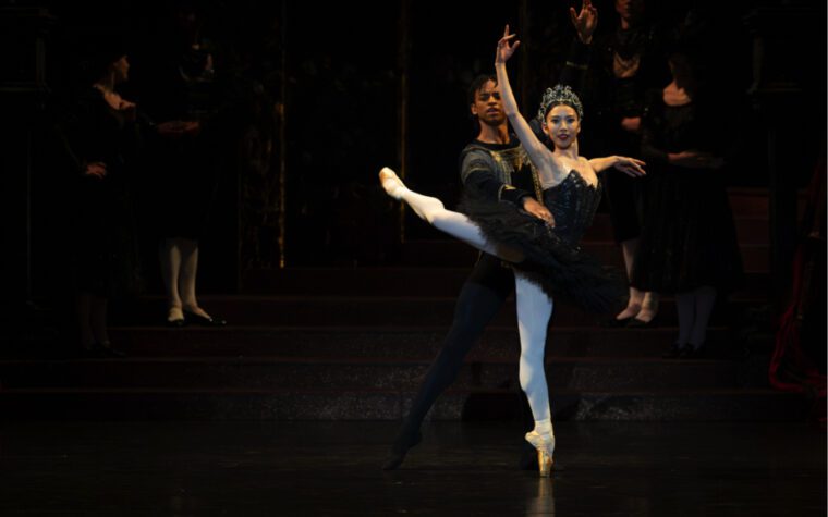 Swan Lake, Birmingham Royal Ballet the ballerina in a black tutu and glittering tiara stands on pointe with her right leg in attitude behind, right arm above her head.  There's a mocking look on her face.  She is supported by the black-clad male dancer who stands behind her.