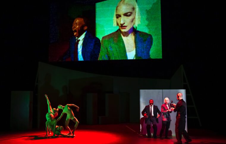 Jess & Morgs Deepfake Multiple images vie for attention. On the high screen close ups of the interviewer and an anguished PM. At stage level on our left three green figures dance. To our right is the full length real time event portrayed on the screen
