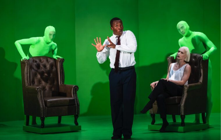 Jess & Morgs Deepfake Against a green background, two figures dressed top to toe in green hold two leather armchairs. On the right sits the female interviewer. The Prime Minister character stands in the center